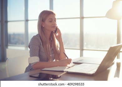 Side view portrait of young businesswoman having business call in office, her workplace, writing down some information. Woman talking on mobile phone, asking questions, looking at pc screen. - Powered by Shutterstock