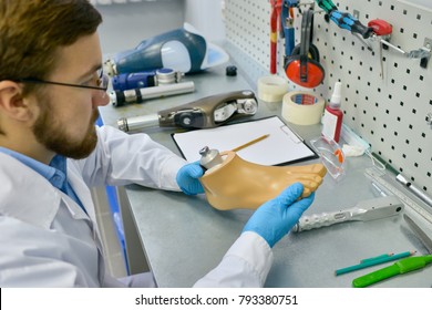 Side View Portrait Of Young Doctor Holding Artificial Foot At Desk In Office, Checking It For Quality And Making Adjustments