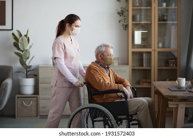 Side View Portrait Of Young Female Nurse Assisting Senior Man In Wheelchair At Retirement Home, Both Wearing Masks, Copy Space