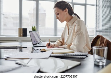 Side View Portrait Of Young Female Boss Taking Notes While Working At Desk In Office Interior