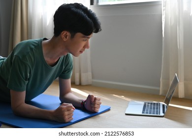 A Side View Portrait Of A Young Handsome Asian Man Planking On A Mat In A Living Room Looking At The Laptop In Front Of Him, For Home Exercise, Online Class Concept.