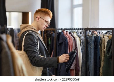 Side view portrait of young man looking at clothes while shopping sustainably in thrift store - Powered by Shutterstock