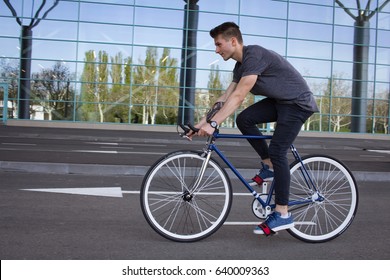 Side view portrait of a young man riding on bicycle in city street. Man on blue bicycle with white wheels, big mirror windows background