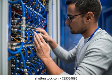 Side view portrait of young man connecting wires in server cabinet while working with supercomputer in data center