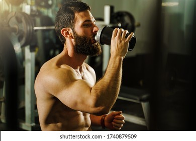 Side View Portrait Of Young Muscular Caucasian Man Bodybuilder Shirtless Male Sitting In Dark Gym Holding Protein Supplement Shaker Drinking Supplementation In Training Waist Up Black Hair And Beard