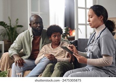 Side view portrait of young nurse consulting African American father and son during at home visit and holding clipboard copy space - Powered by Shutterstock