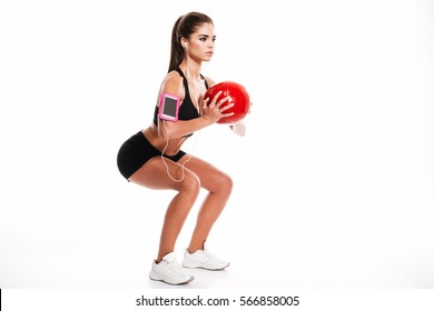 Side view portrait of a young pretty sportswoman doing squats with heavy fitness ball and listening to music isolated on a white background