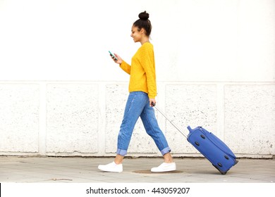 Side view portrait of young woman walking with travel bag and using mobile phone