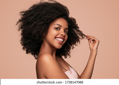 Seitenansicht eines positiven afroamerikanischen Modells, das nach Kamera lächelt und sauberes, lockiges Haar nach hygienischer Routine vor braunem Hintergrund berührt