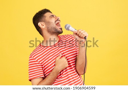 Side view positive bearded man in striped t-shirt loudly singing song holding microphone in hand, having fun resting in karaoke, singer performance. Indoor studio shot isolated on yellow background
