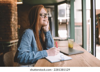 Side view positive female in eyeglasses sitting and hand leaning   touching chin at table and laptop juice glass   taking notes in notebook while looking away at street in daylight in cafe