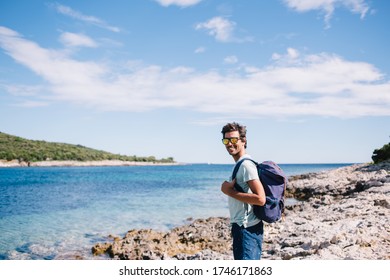 Side view of positive man with backpack smiling and looking at camera while standing on stony coast against sea and cloudy sky