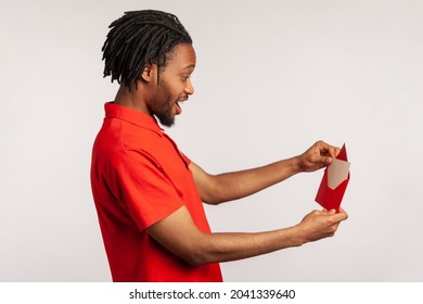 Side View Of Positive Man With Dreadlocks Wearing Red Casual Style T-shirt, Opening Letter In Red Envelope, Holding Greeting Card And Feels Happy. Indoor Studio Shot Isolated On Gray Background.