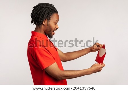 Side view of positive man with dreadlocks wearing red casual style T-shirt, opening letter in red envelope, holding greeting card and feels happy. Indoor studio shot isolated on gray background.