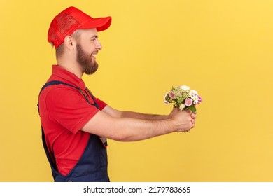 Side View Of Positive Satisfied Bearded Delivery Man Giving Bouquet Of Flowers, Bringing Order, Looking At Camera With Happy Expression. Indoor Studio Shot Isolated On Yellow Background.