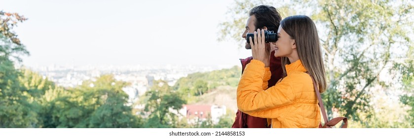 Side view of positive traveler looking through binoculars near boyfriend outdoors, banner