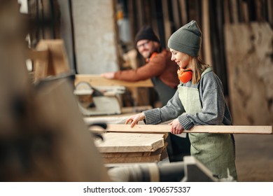 Side view of positive young female woodworker in apron working with lumber in carpentry workshop with colleague
