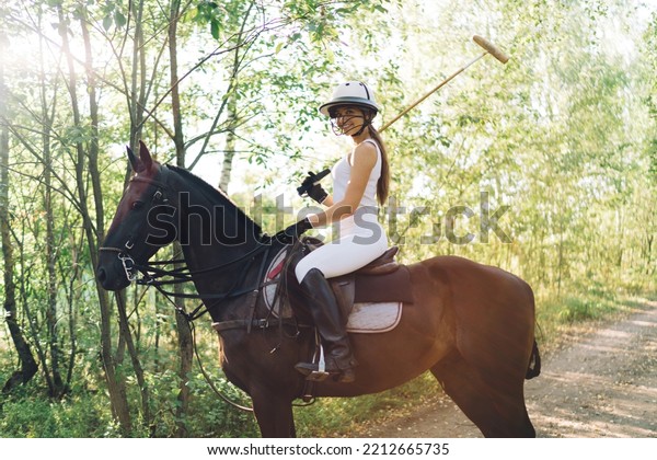 Side view of positive\
young female equestrian in riding hat boots and breeches smiling\
and looking at camera while sitting on horse with polo mallet in\
hand before training