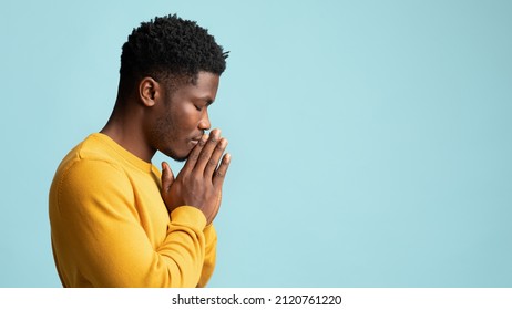 Side view of praying african american young man in yellow, holding palms by his lips and whispering pray, asking for grace over blue studio background, panorama wuth copy space