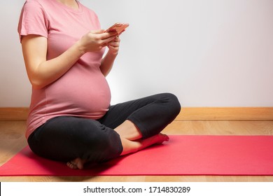 Side View Of Pregnant Young Woman In Pink T-shirt And Black Pants Using Mobile Phone Sitting On Yoga Mat