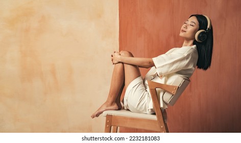 Side view of pretty asian girl sitting on chair at studio enjoying favorite songs or classical music in headphones on brown background with copy space - Shutterstock ID 2232181083