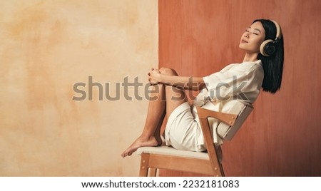 Side view of pretty asian girl sitting on chair at studio enjoying favorite songs or classical music in headphones on brown background with copy space