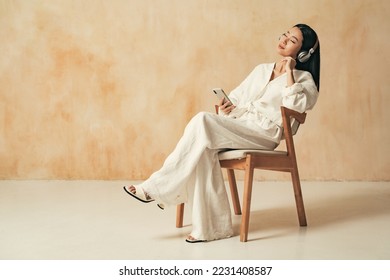 Side view of pretty asian woman sitting on chair on brown copy space background in home clothes, listening music in wireless headphones with closed eyes, using audio player app for phone