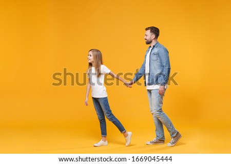 Side view of pretty bearded man in casual clothes have fun with child baby girl. Father little kid daughter isolated on yellow wall background. Love family parenthood childhood concept. Holding hands