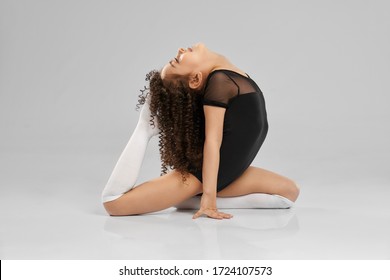 Side view of pretty girl in black sportswear and white knee socks posing on floor, isolated on gray studio background. Little female professional gymnast with curly hair showing flexibility, training.