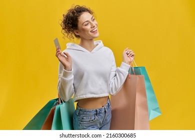 Side View Of Pretty Girl Holding Packages And Discount Card. Young Female With Curly Blonde Hair Doing Shopping, Looking Forward, Smiling. Concept Of Youth And Modern Culture.