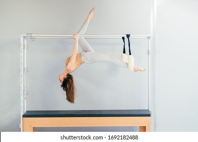 Side View Of Pretty Pilates Instructor Hanging Upside Down From Trapeze Table In Studio Over Grey Wall Background. Pilates, Fitness, Sport, Training And People Concept