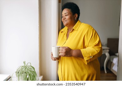 Side view of pretty senior african american female in yellow shirt holding white cup of herbal tea standing near window, looking through admiring city life, having rest and relaxing - Powered by Shutterstock