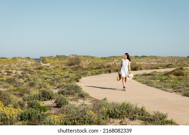 Side view of pretty woman holding handbag and straw hat while walking on pathway on beach