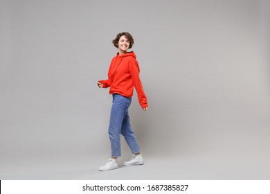Side view of pretty young brunette woman girl in casual red hoodie, blue jeans posing isolated on grey wall background studio portrait. People lifestyle concept. Mock up copy space. Looking aside