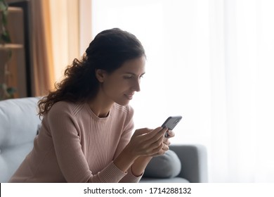Side view pretty young woman sitting on comfortable couch, looking at mobile phone screen, involved in pleasant conversation in social networks or reading interesting article information on website. - Shutterstock ID 1714288132