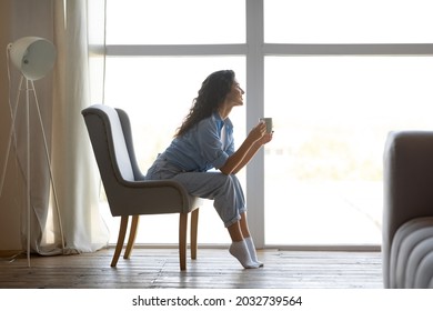 Side View Of Pretty Young Woman Drinking Hot Coffee In Cozy Chair Near Window Indoors, Copy Space. Peaceful Millennial Lady Having Relaxing Day, Chilling On Lazy Morning Indoors