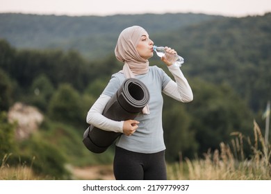 Side view of pretty young woman in hijab and activewear drinking water from bottle while standing at green park. Muslim female relaxing and refreshing after yoga practice outdoors. - Powered by Shutterstock