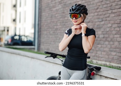 Side View Of Pretty Young Woman Wearing Safety Helmet And Mirrored Glasses While Standing On Street With Black Bike. Female Cyclist Preparing For Outdoors Workout.