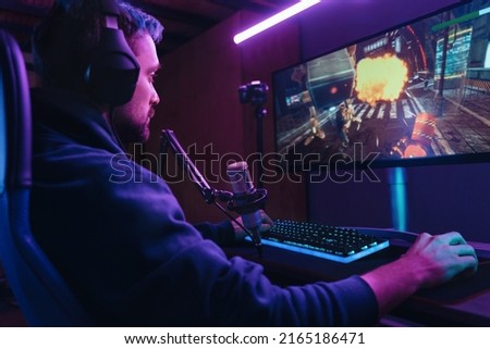 Side view of pro streamer playing video game competition use professional gaming setup. Gamer sitting on gaming chair using professional headphones and microphone late at night at home. Cyber sport