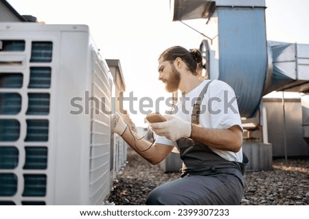 Side view of professional builder sitting in front of air conditioner and using measuring device on roof of factory. Caucasian focused male with braided hair testing voltage of climate control unit.