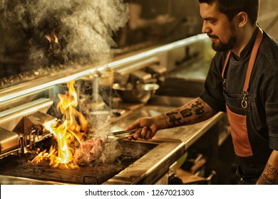 Side View Of Professional Chef Cooking Delicious Juicy Beef Steak On Flaming Grill. Handsome Man With Beard And Tattoos On Hand Preparing Food In Modern Restaurant Kitchen. 