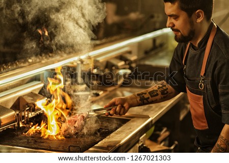 Side view of professional chef cooking delicious juicy beef steak on flaming grill. Handsome man with beard and tattoos on hand preparing food in modern restaurant kitchen. 
