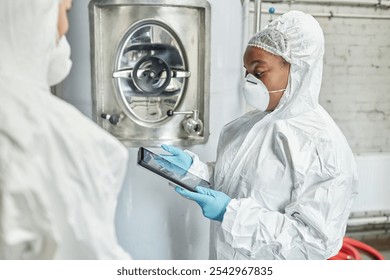 Side view of professional female African American supervisor in mask and hazmat suit using tablet computer while discussing with colleague production process at food factory, copy space - Powered by Shutterstock