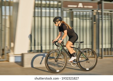 Side view of professional female cyclist in black cycling garment and protective gear riding bicycle in city, rushing and passing outdoors on a sunny day