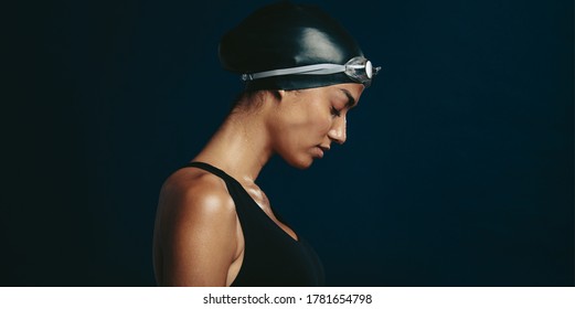 Side view of a professional female swimmer with goggles and a hat. Woman wearing black swimsuit, swimming cap and goggles on dark background looking exhausted.