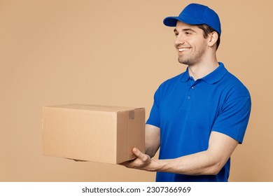Side view professional happy delivery guy employee man wear blue cap t-shirt uniform workwear work as dealer courier hold give cardboard box isolated on plain light beige background. Service concept
