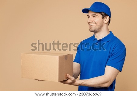 Side view professional happy delivery guy employee man wear blue cap t-shirt uniform workwear work as dealer courier hold give cardboard box isolated on plain light beige background. Service concept