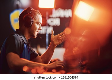 Side view of a professional male cybersport gamer wearing headphones talking by microphone with team member while playing online video game