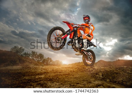 Side View of the Professional Motorcycle Rider Driving on the mountains and Further Down the Off-Road Track. It's Sunset and Track is Covered with Smoke Mist.