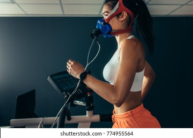 Side View Of Professional Sportswoman With Mask Running On Treadmill In Gym. Female Athlete In Sports Science Lab Measuring Her Performance And Oxygen Consumption.